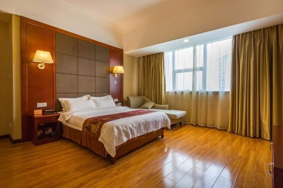 A hotel room with a large bed, a desk, a chair, a window with curtains, and a wall-mounted lamp.
