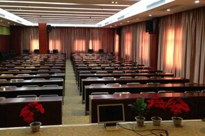 Un salón de conferencias con filas de sillas, un escenario con ponentes y cortinas en las paredes.