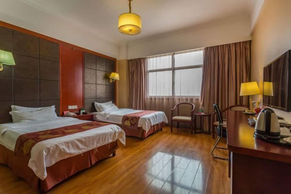 A hotel room with two beds, a desk, a chair, a large window with curtains, and a hanging light fixture.
