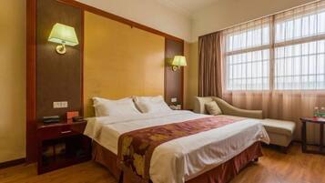 Een hotelkamer met een bed, bedlampen, een stoel, een klein tafeltje en een raam met gordijnen.