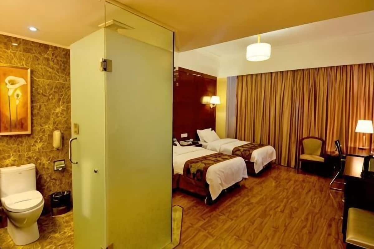 A hotel room with two beds, a bathroom with a toilet, a desk with a chair, and a large window with curtains.