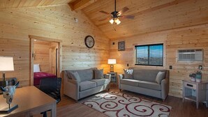 L’intérieur d’un chalet confortable, avec deux canapés, une table à manger, un ventilateur de plafond et une fenêtre équipée de stores.