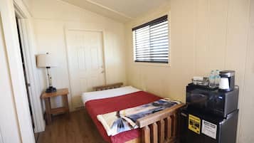 A bedroom with a bed, a nightstand, a lamp, a window with blinds, and a refrigerator.