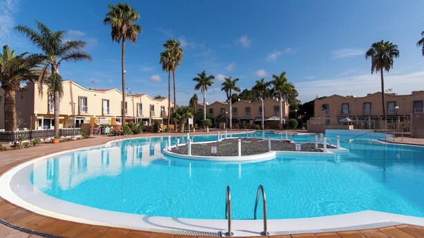 A large, clear swimming pool surrounded by palm trees and a row of beige buildings with balconies. - Home2Book Green Oasis Charming Duplex Maspalomas (Maspalomas)