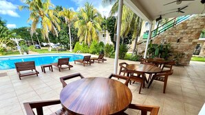 A covered outdoor seating area with wooden tables and chairs, a pool in the background, and palm trees. - Villas Paraiso (Mérida)