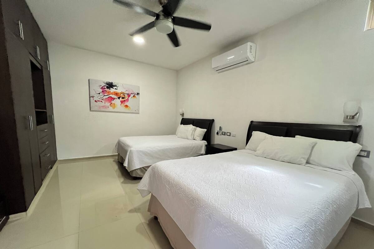 Habitación de hotel con dos camas, ventilador de techo, unidad de aire acondicionado, un cuadro en la pared y un armario de madera.