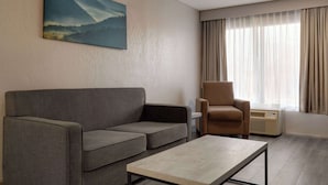 A modern living room with a grey sofa, a brown armchair, and a small table. A painting of a mountain landscape hangs on the wall. - Baymont By Wyndham Emporia Va (Emporia)