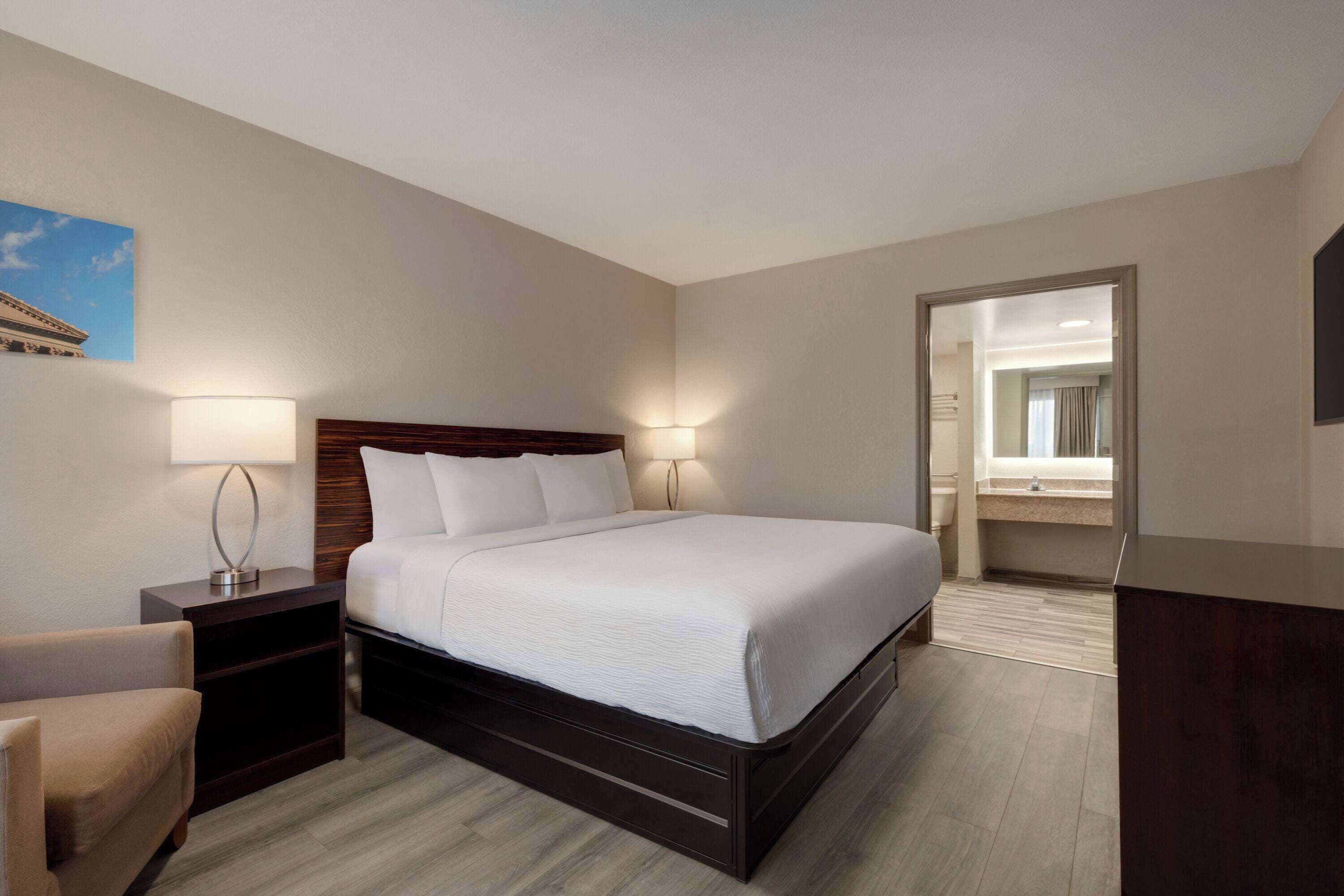 Habitación de hotel con una cama grande, lámparas de noche, una silla, un escritorio y un baño visible a través de una puerta abierta.