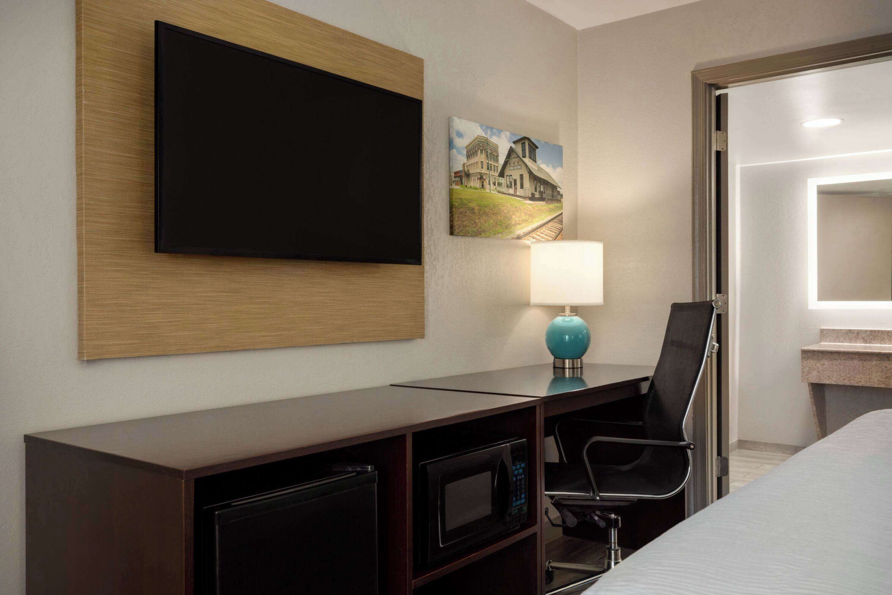 Une chambre d’hôtel équipée d’un téléviseur à écran plat fixé au mur, d’un bureau avec une lampe, d’un four à micro-ondes et d’une chaise.