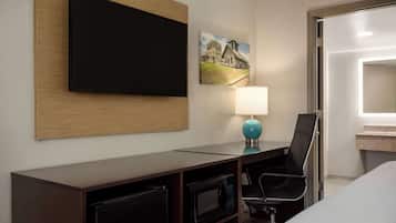 Une chambre d’hôtel équipée d’un téléviseur à écran plat fixé au mur, d’un bureau avec une lampe, d’un four à micro-ondes et d’une chaise.