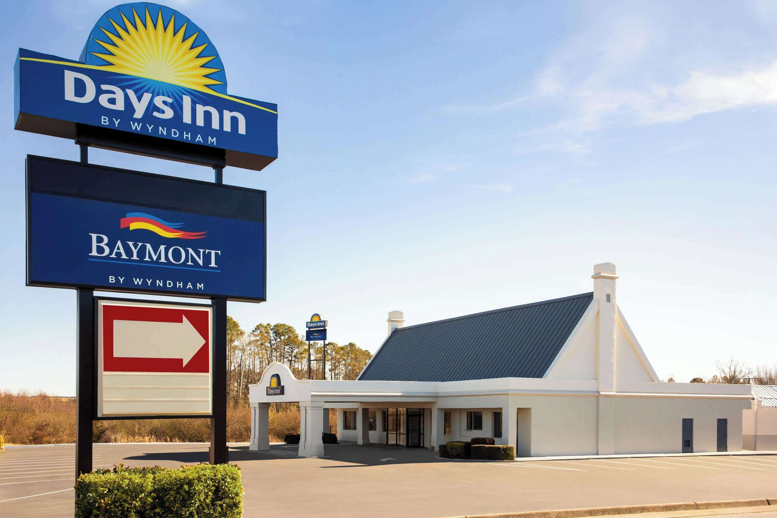 Señal de Days Inn by Wyndham Baymont con flecha indicadora, estacionamiento y un edificio de fachada blanca y techo oscuro.