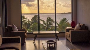 A modern living room with a large window overlooking a body of water and palm trees. - Ramada Suites By Wyndham Seafront Coral Coast (Korotogo)