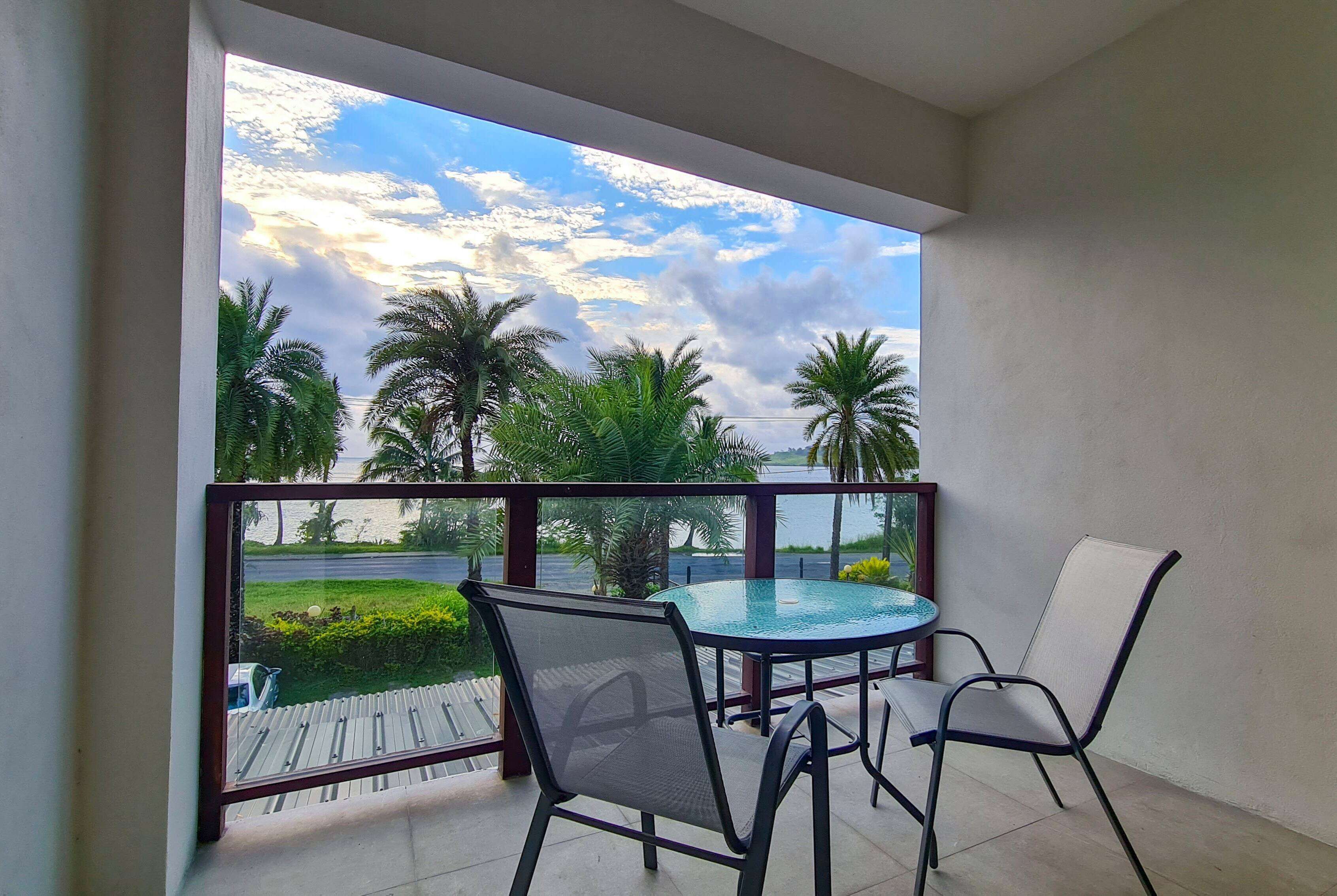 A balcony with a glass table and chairs, overlooking a palm tree-lined street and a body of water.