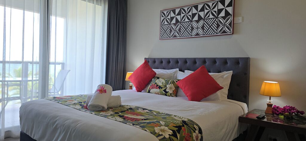 A hotel room with a large bed, a nightstand with a lamp, a decorative wall piece, and a view of the outdoors.