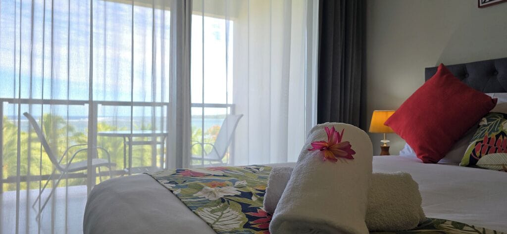 Habitación de hotel con balcón, cama con colcha estampada y almohada roja.