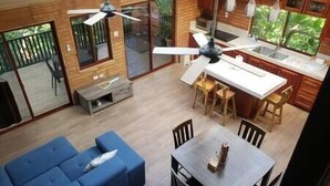 A spacious room with a blue sofa, a dining table with chairs, a ceiling fan, and large windows offering a view of greenery outside.
