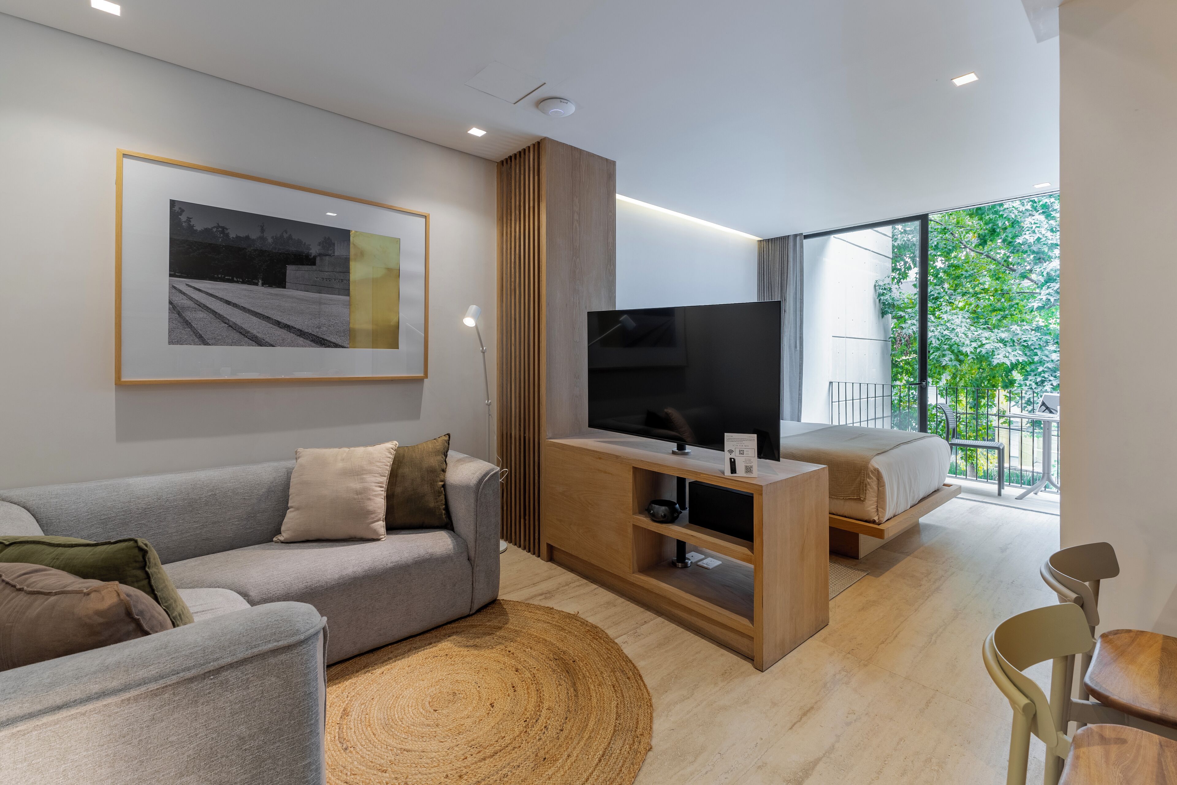 A modern living room with a grey sofa, a wooden TV stand, a bed with a white mattress, and a large window offering a view of greenery.