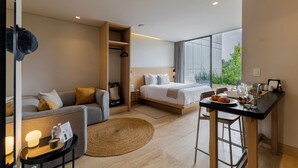 A modern hotel room with a sofa, a bed, a dining table, and a large window offering a view of greenery.