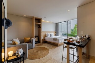 Habitación de hotel moderna con sofá, cama, mesa de comedor y un gran ventanal con vistas a zonas verdes.