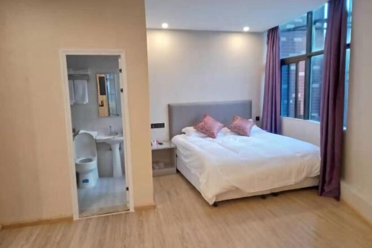 A modern hotel room with a large bed, a nightstand, a window with curtains, and a bathroom visible through an open door.