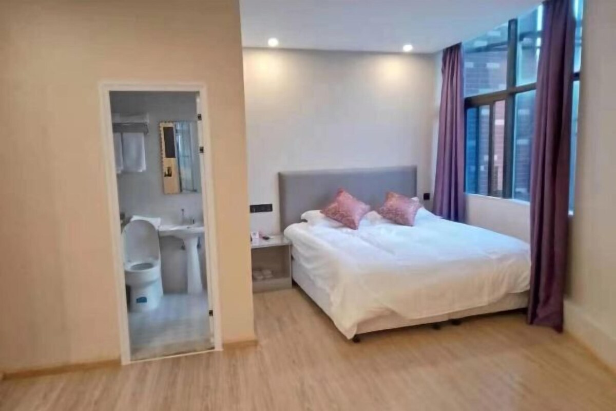 A hotel room with a bed, a nightstand, a window with curtains, and a bathroom visible through an open door.