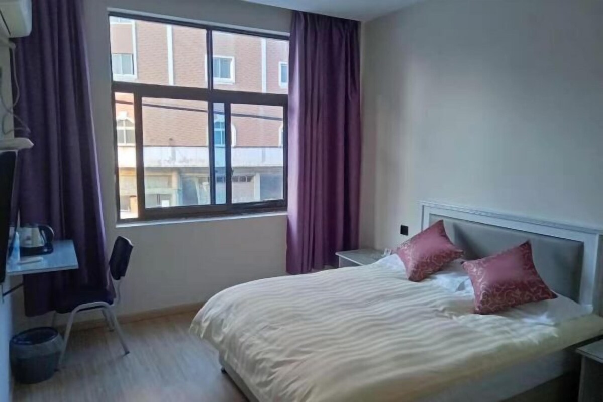 A hotel room with a bed, a desk with a chair, a window, and a view of buildings outside.