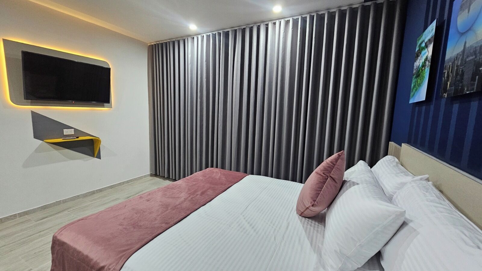 Una habitación de hotel moderna con una cama grande, un televisor de pantalla plana montado en la pared y un cuadro con un paisaje urbano.