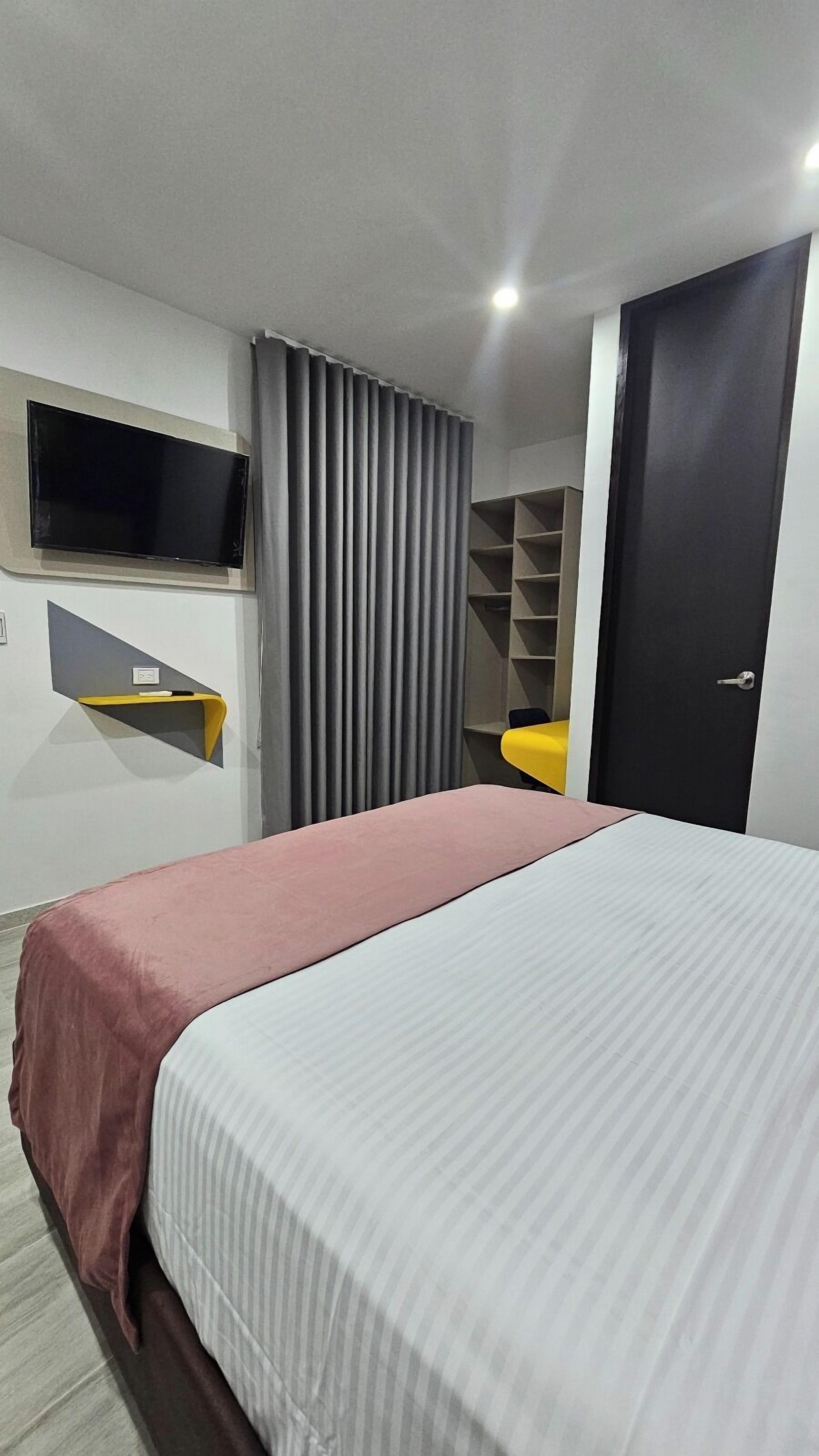 A modern hotel room with a bed, a TV mounted on the wall, a desk with a chair, and a wardrobe.
