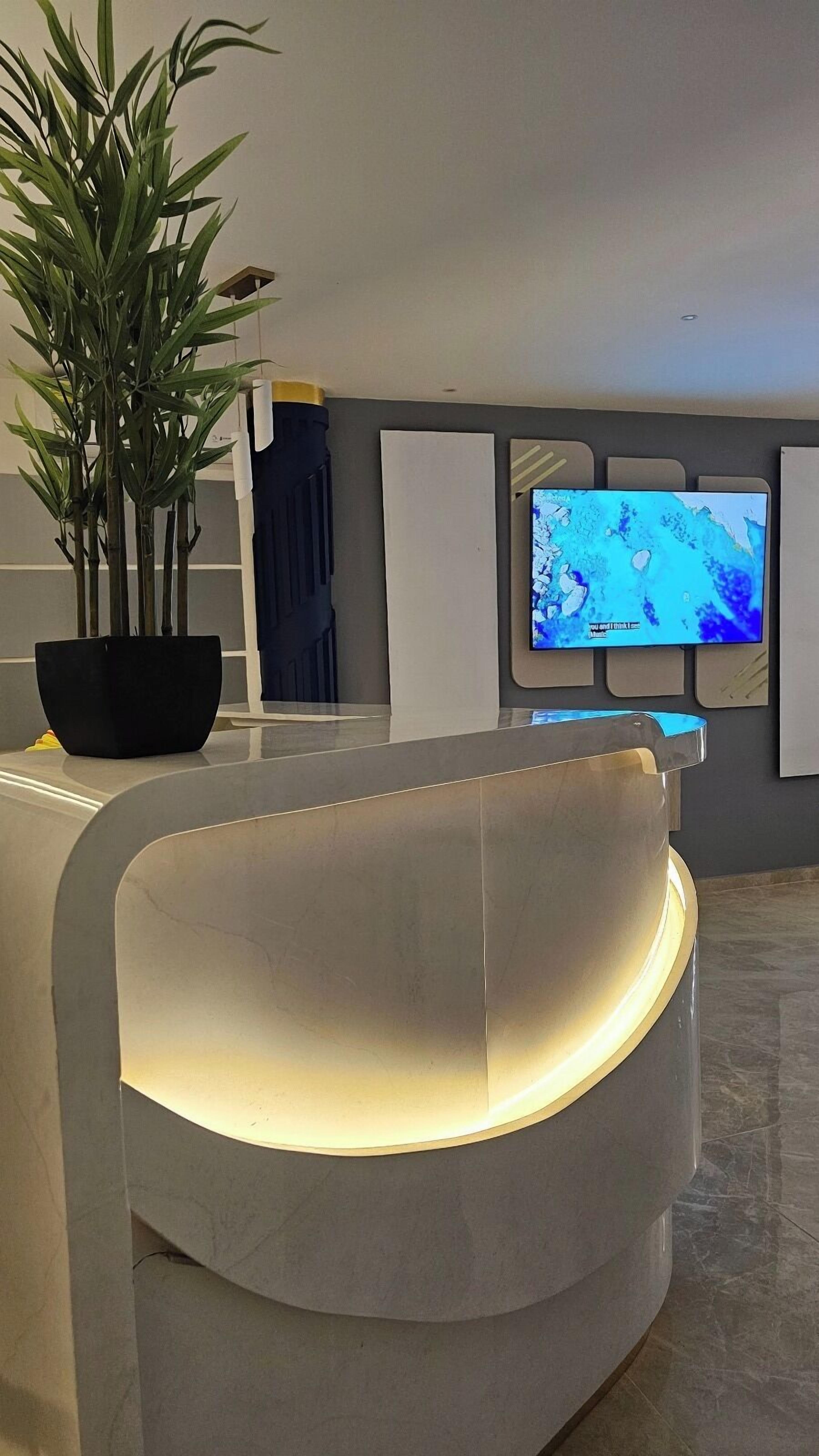 A modern reception area with a curved white reception desk, a large potted plant, and a flat-screen TV mounted on the wall.