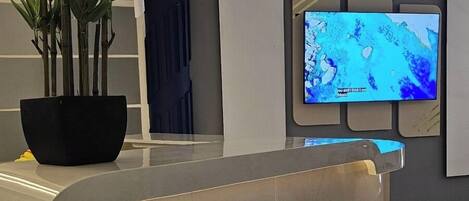 Un área de recepción moderna con un mostrador blanco curvo, una planta grande en maceta y un televisor de pantalla plana montado en la pared.