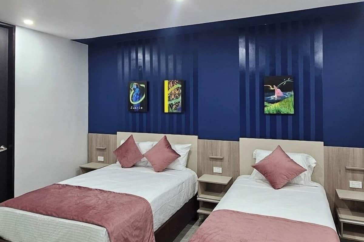 Une chambre d’hôtel avec deux lits, chacun doté d’un cadre en bois et de draps blancs. Il y a deux tables de chevet avec des lampes, et les murs sont peints en bleu et ornés de trois tableaux encadrés.