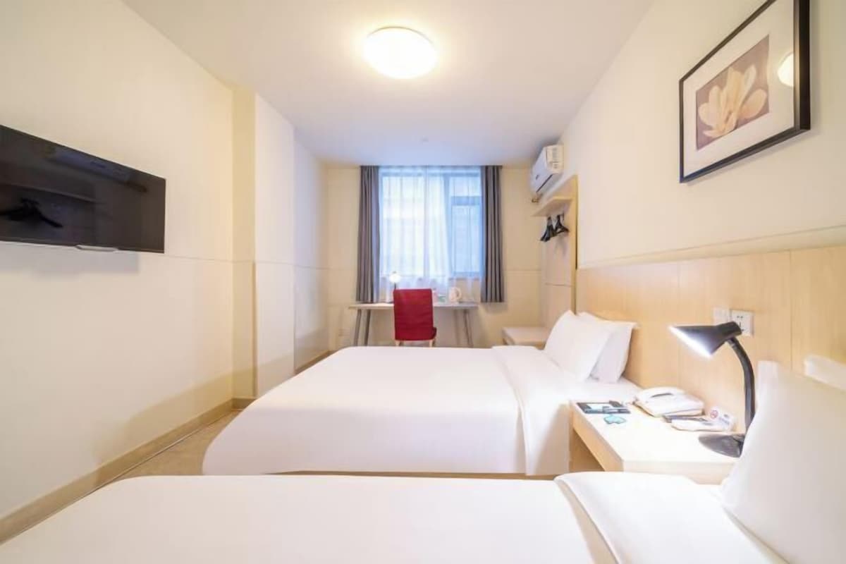 A hotel room with two beds, a desk with a chair, a TV mounted on the wall, and a window with curtains.
