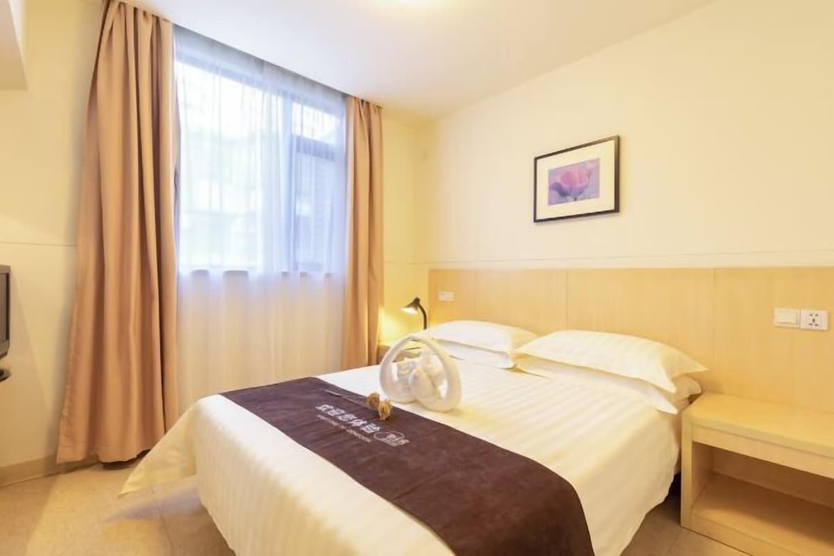 A hotel room with a bed, a bedside table, a lamp, a picture on the wall, and a window with curtains.