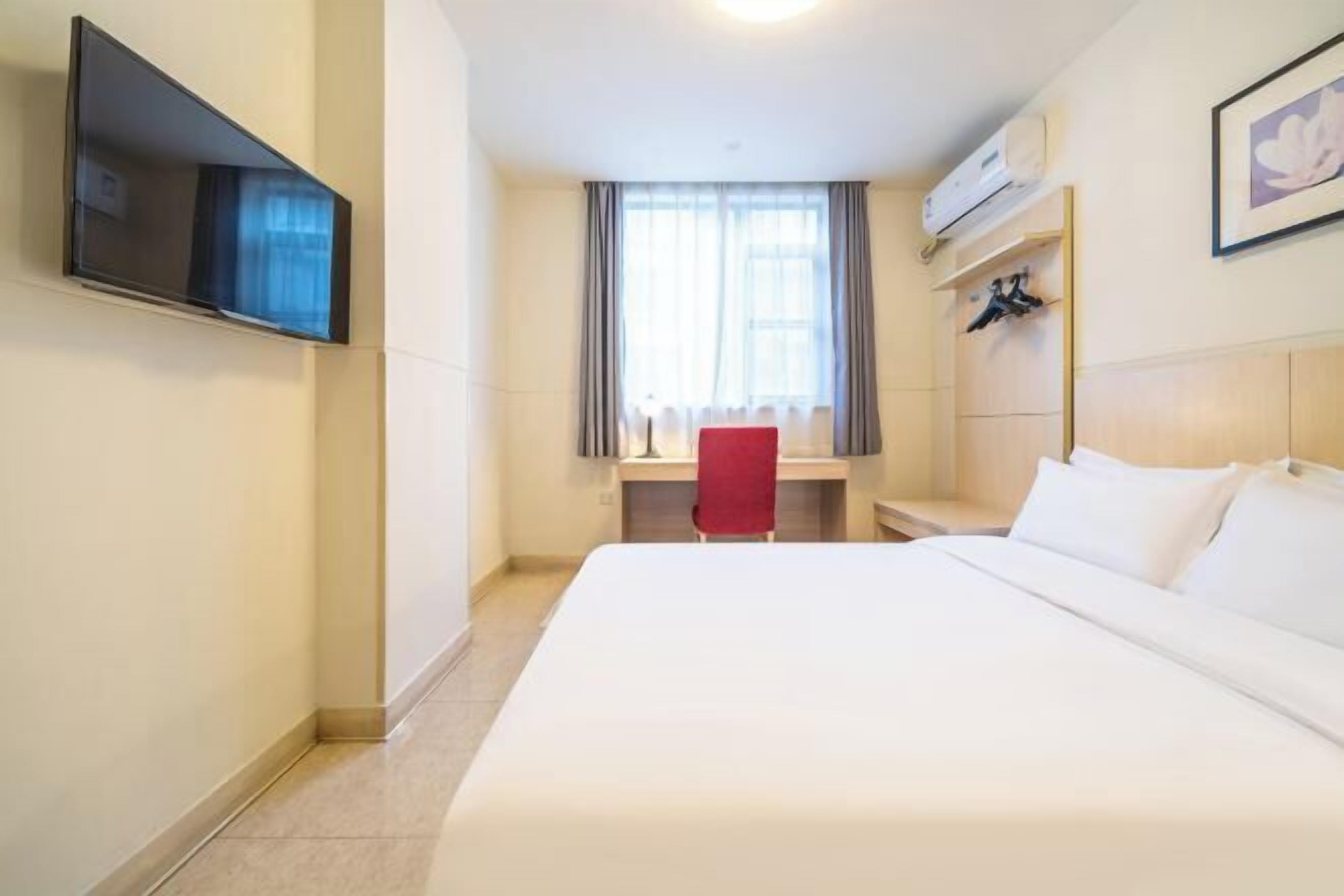 A hotel room with a large bed, a desk with a chair, a TV mounted on the wall, and a window with curtains.