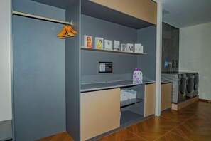 Zona de lavandería con lavadora, secadora y estantes con ropa doblada y útiles de limpieza.