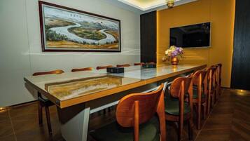 A dining area with a long table, chairs, a painting, a vase with flowers, and a television on the wall.