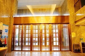 Aula, jossa on kaksinkertaiset ovet, vastaanottotiski ja seinälle kiinnitetty informaatiotaulu.