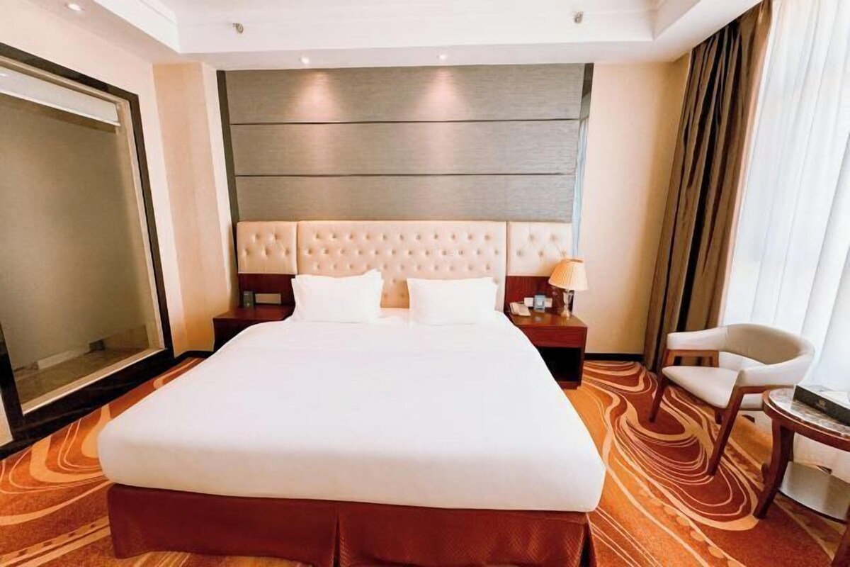 Quarto de hotel com uma cama grande, cabeceira, mesa de cabeceira, uma cadeira e um espelho grande.
