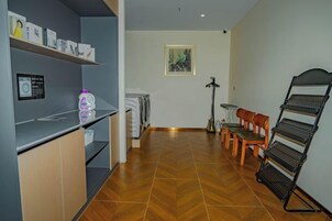 Habitación de hotel con kitchenette, zona de comedor con sillas, un estante metálico negro y un cuadro en la pared.