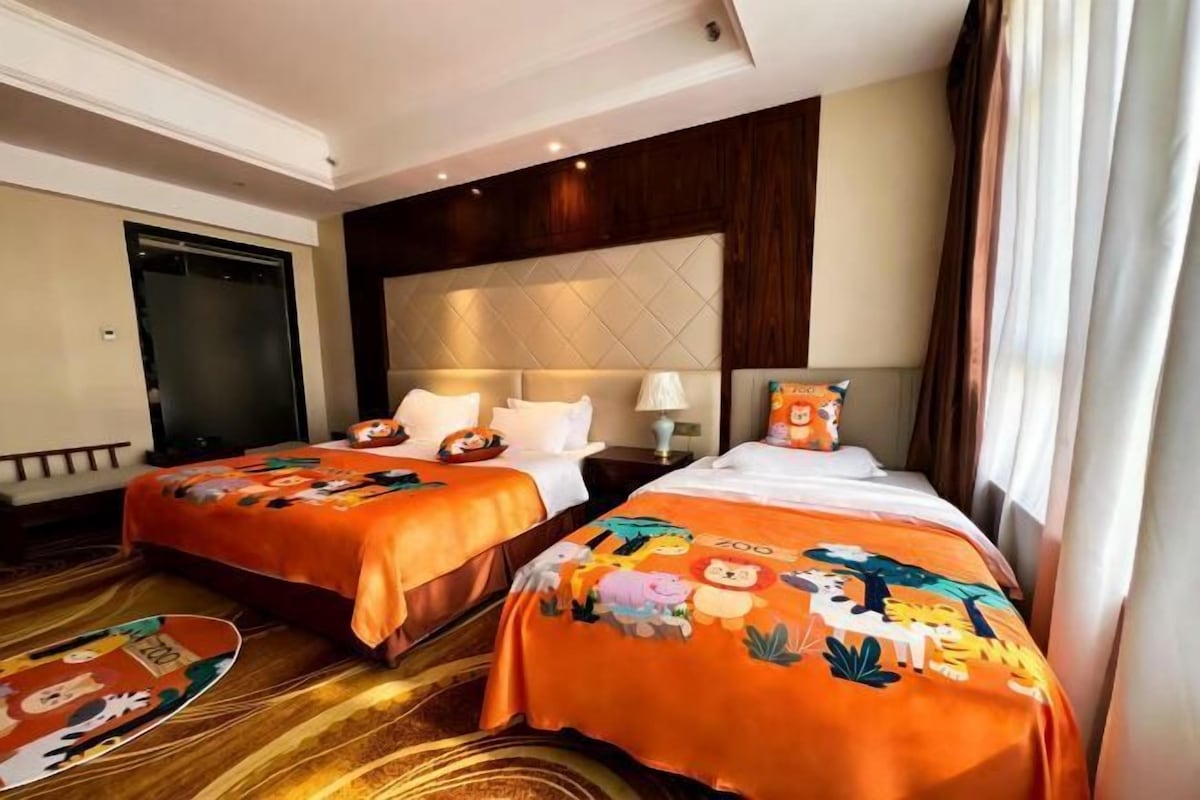 Quarto de hotel com duas camas, cada uma com colcha laranja com personagens de desenho animado. Há uma janela grande com cortinas, piso de madeira e cabeceira com padrão acolchoado.