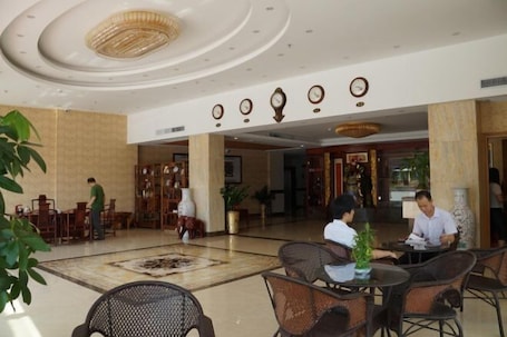 A spacious lobby with a high ceiling, a central chandelier, and a clock on the wall. There are several tables and chairs arranged for guests.