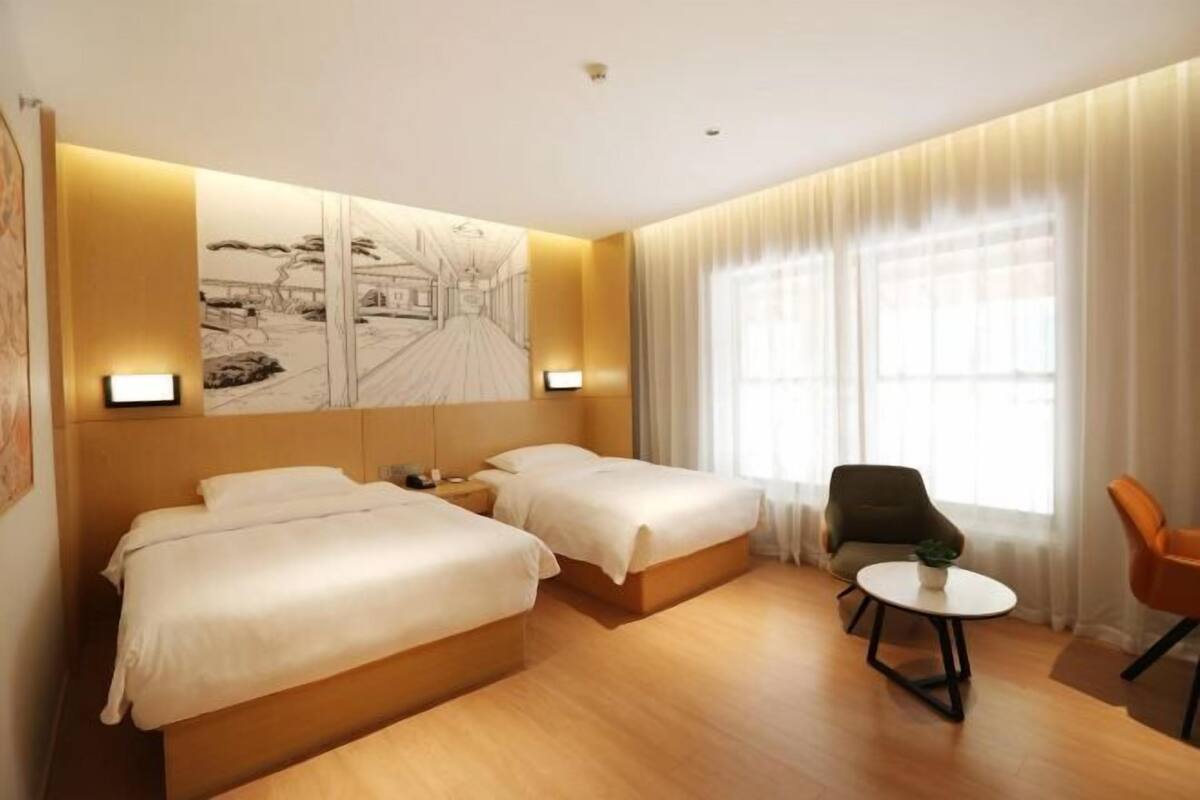 Une chambre d’hôtel avec deux lits, une petite table, une chaise et une grande fenêtre avec des rideaux.