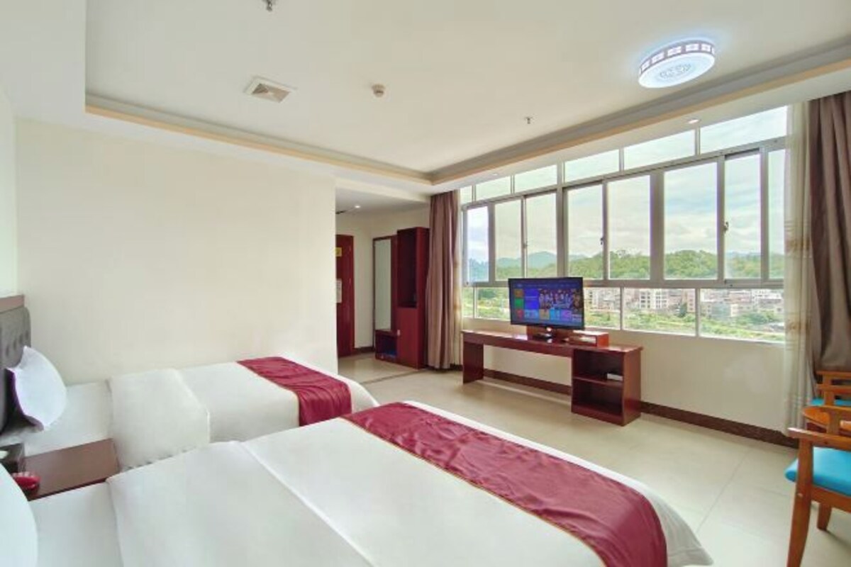 A hotel room with two beds, a television, a desk, and a large window offering a view of the outdoors.