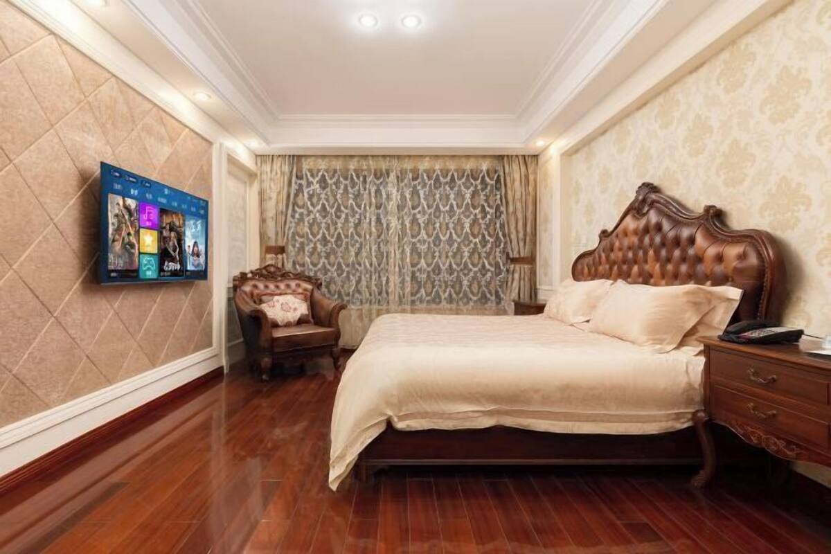 A bedroom with a large bed, a wooden nightstand, a TV mounted on the wall, and a patterned curtain.