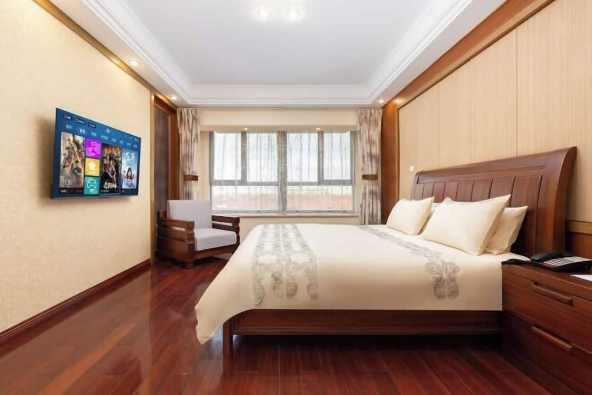 A hotel room with a large bed, a bedside table, a chair, a TV mounted on the wall, and a window with curtains.
