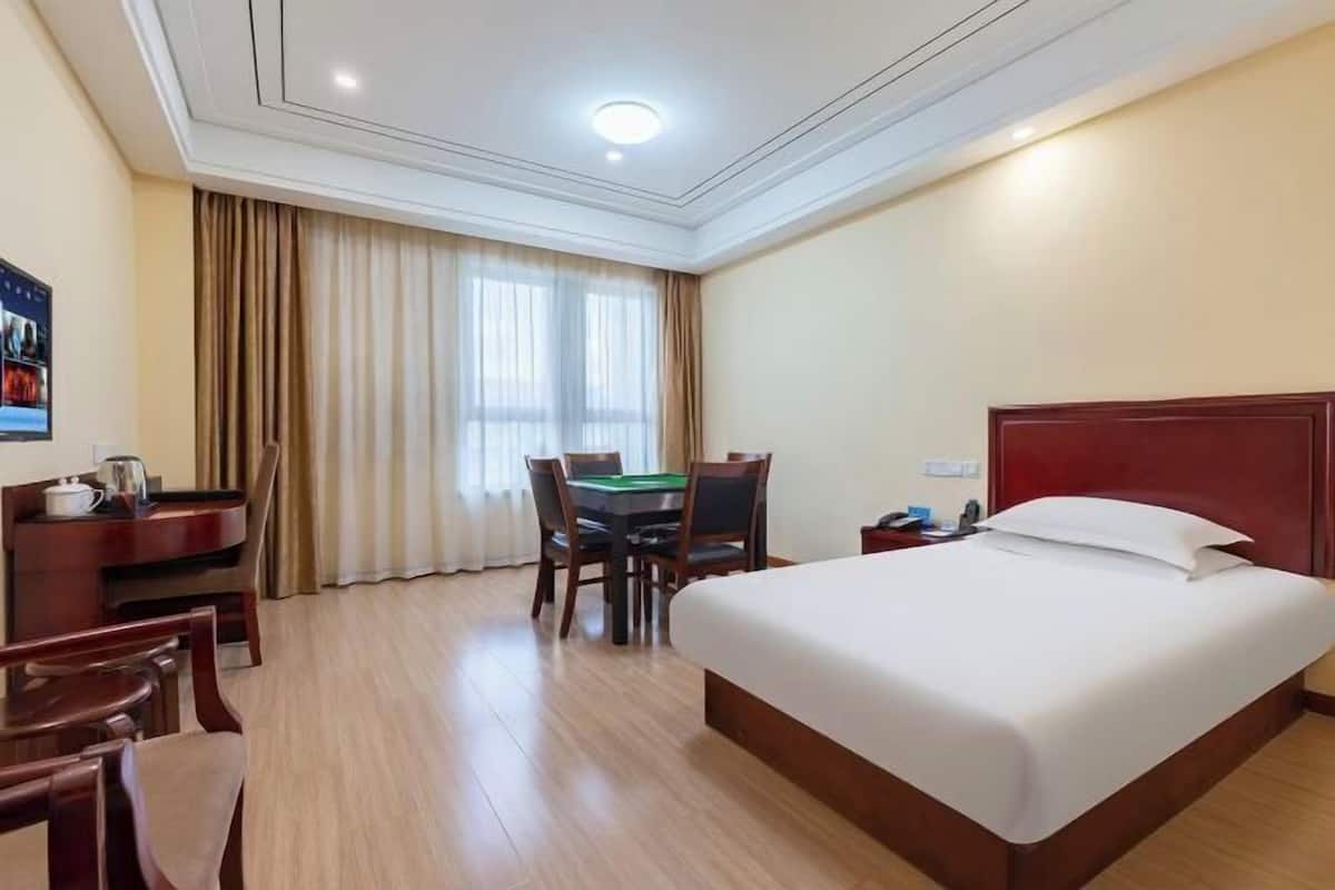 A hotel room with a large bed, a desk with a chair, a table with chairs, and a window with curtains.