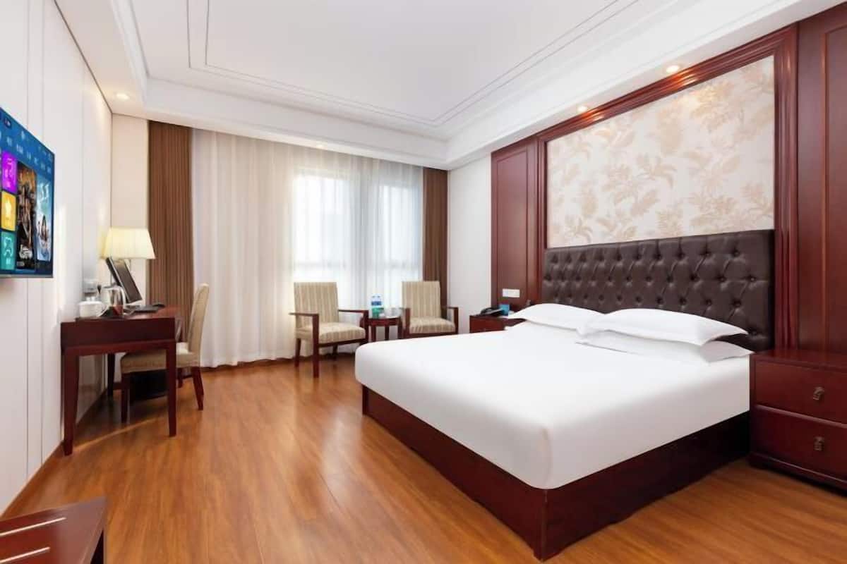 A hotel room with a large bed, a desk with a lamp, a chair, and a small table.