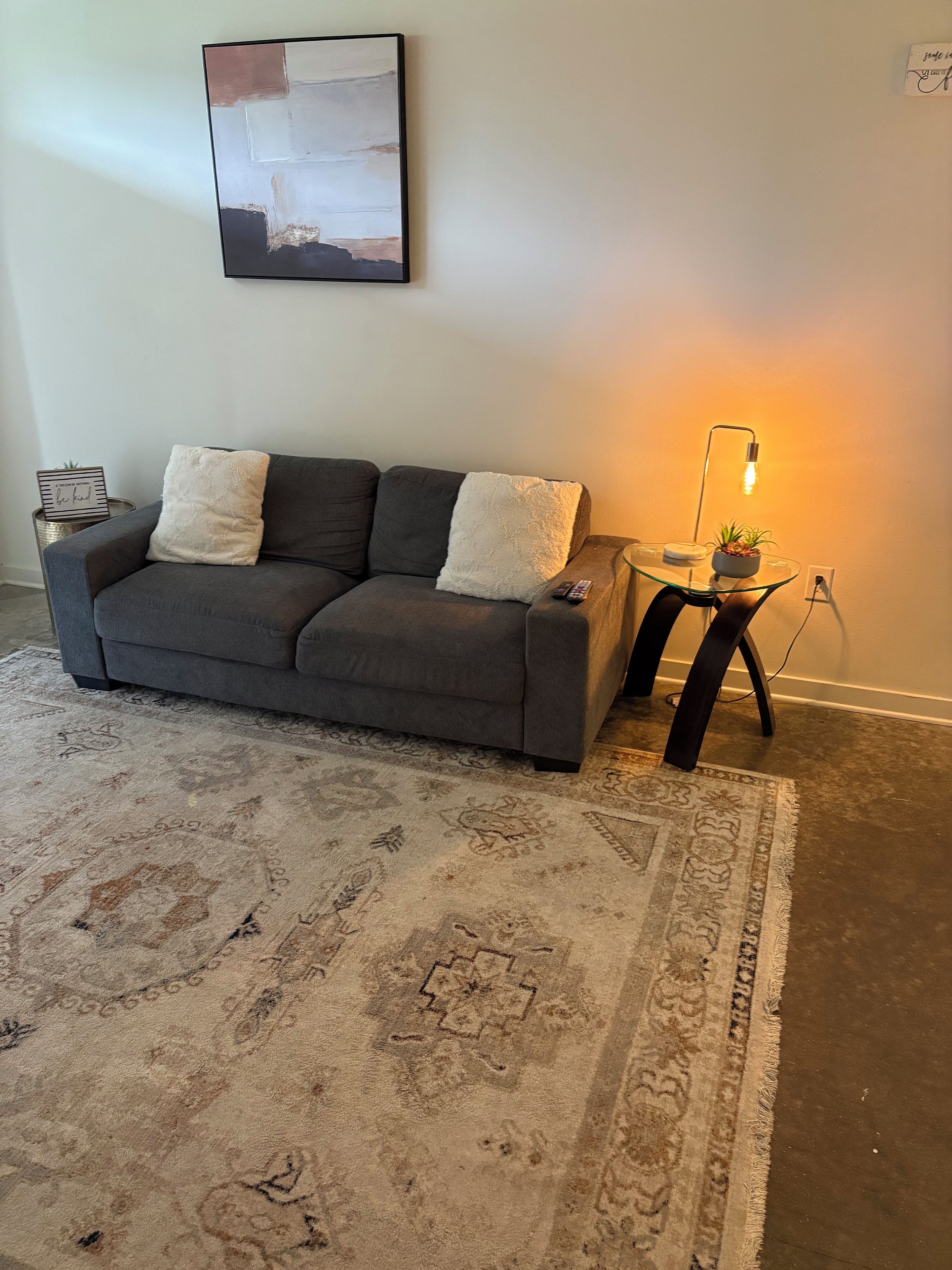 Ein Wohnzimmer mit einem grauen Sofa, einem gemusterten Teppich, einem kleinen Beistelltisch mit einer Lampe und einem an der Wand hängenden Bild.