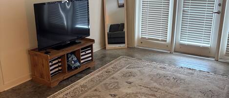 A living room with a flat-screen TV on a wooden stand, a large patterned rug, and a sliding glass door with blinds.