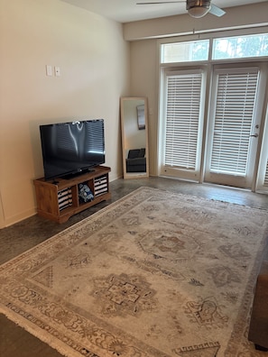 A living room with a flat-screen TV on a wooden stand, a large patterned rug, and a sliding glass door with blinds.